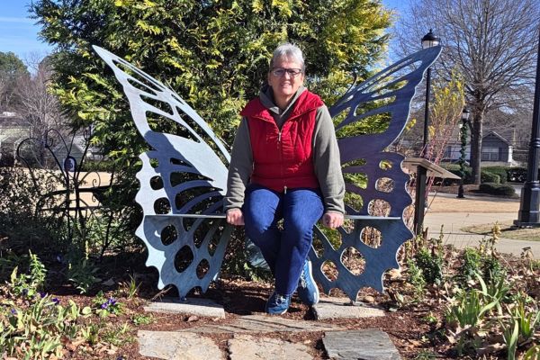 Claudette Eames sitting on a butterfly-shaped bench in a park, surrounded by trees, reflecting quietly on stability and calm at the end of the year.