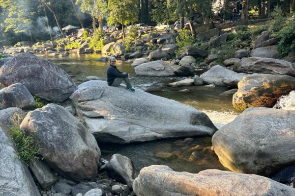 Claudette sitting on a rock beside a flowing brook, reflecting on building a legacy business rooted in wellness, values, and long-term support.