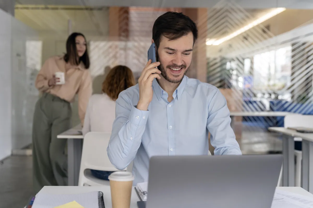 Man using a phone and laptop in an office, representing real-time lead conversion through Pay Per Call and SMS marketing strategies.