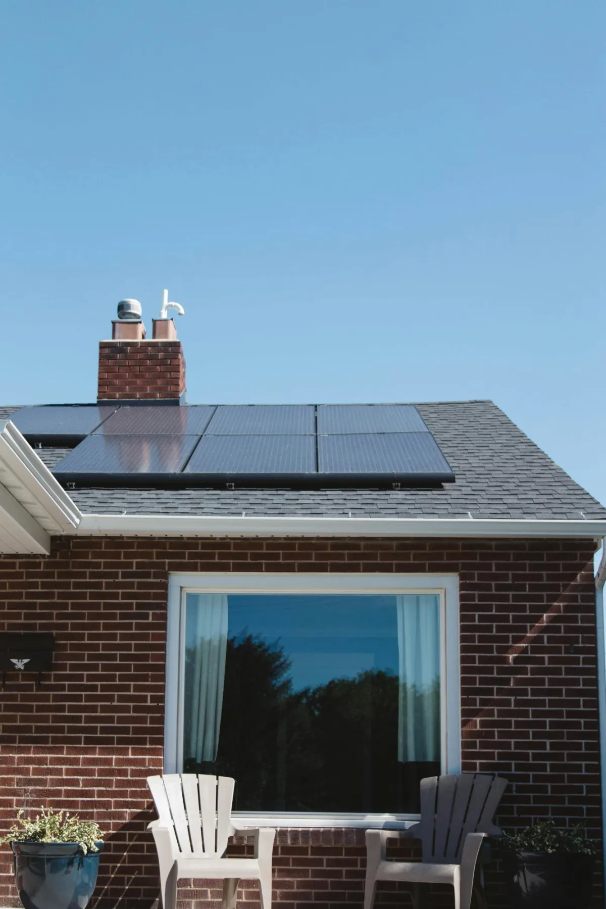 A brick house with solar panels installed on its roof, two patio chairs, and a potted plant in the foreground.