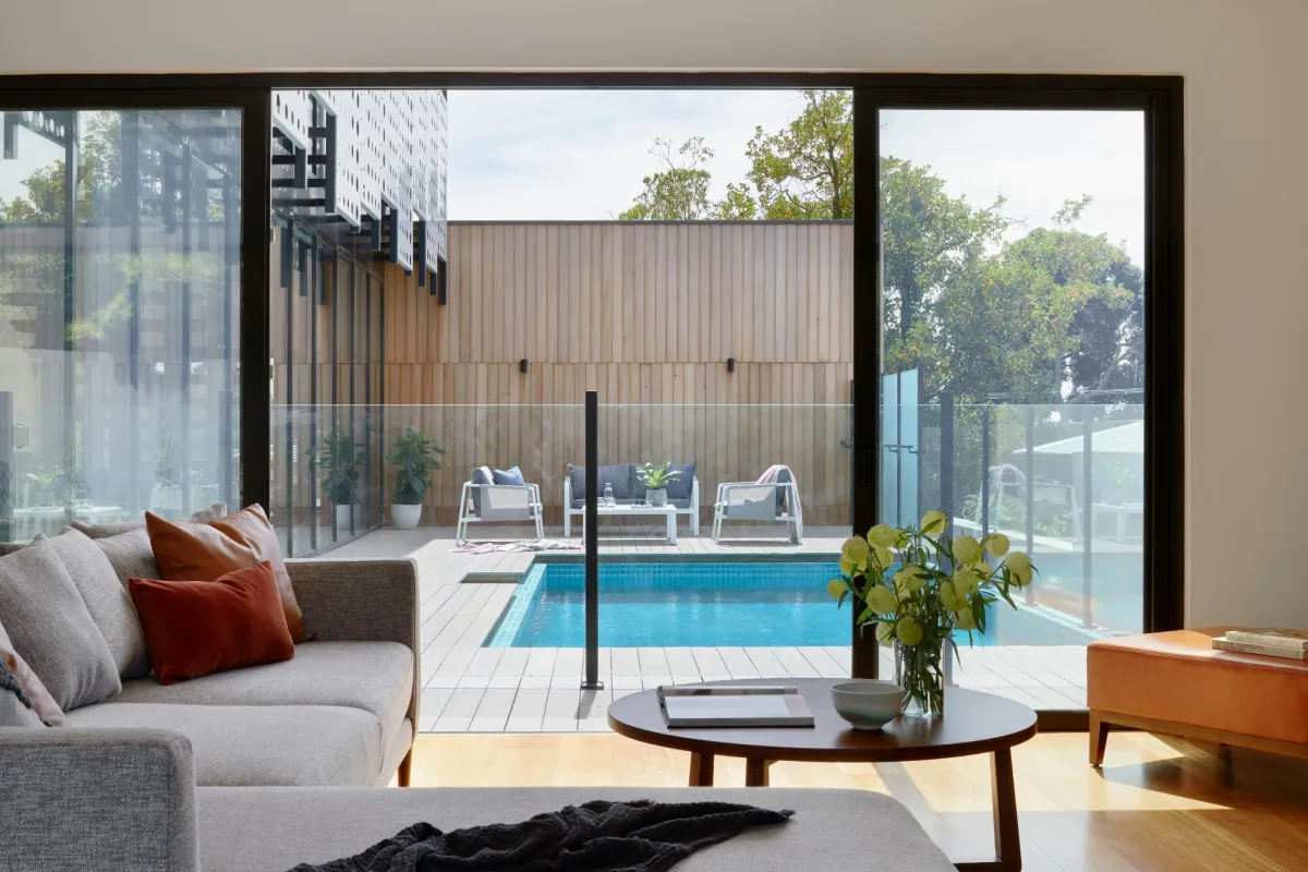 A modern living room with a large sliding glass door opening to a backyard pool and lounge area, featuring a cozy sofa with pillows, a wooden coffee table with a plant, and natural lighting.