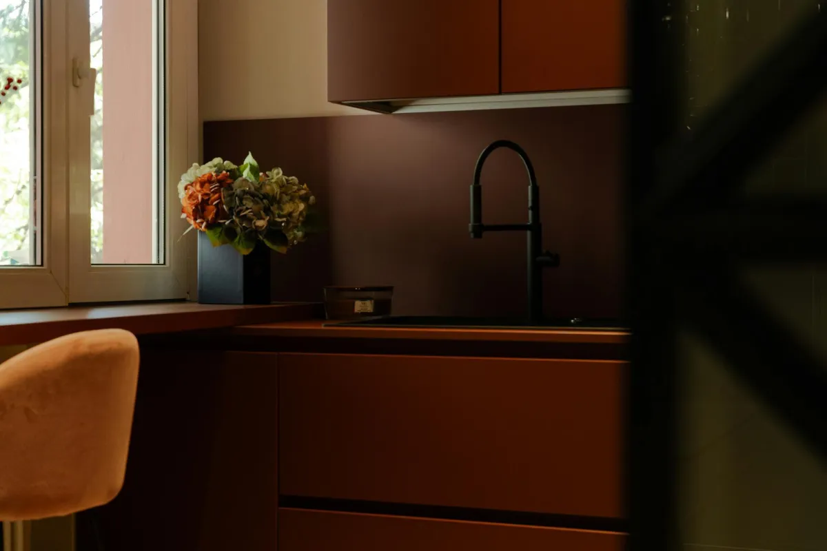 A modern kitchen with warm-toned cabinetry, a black faucet, and soft lighting, featuring a vase with flowers near a window.