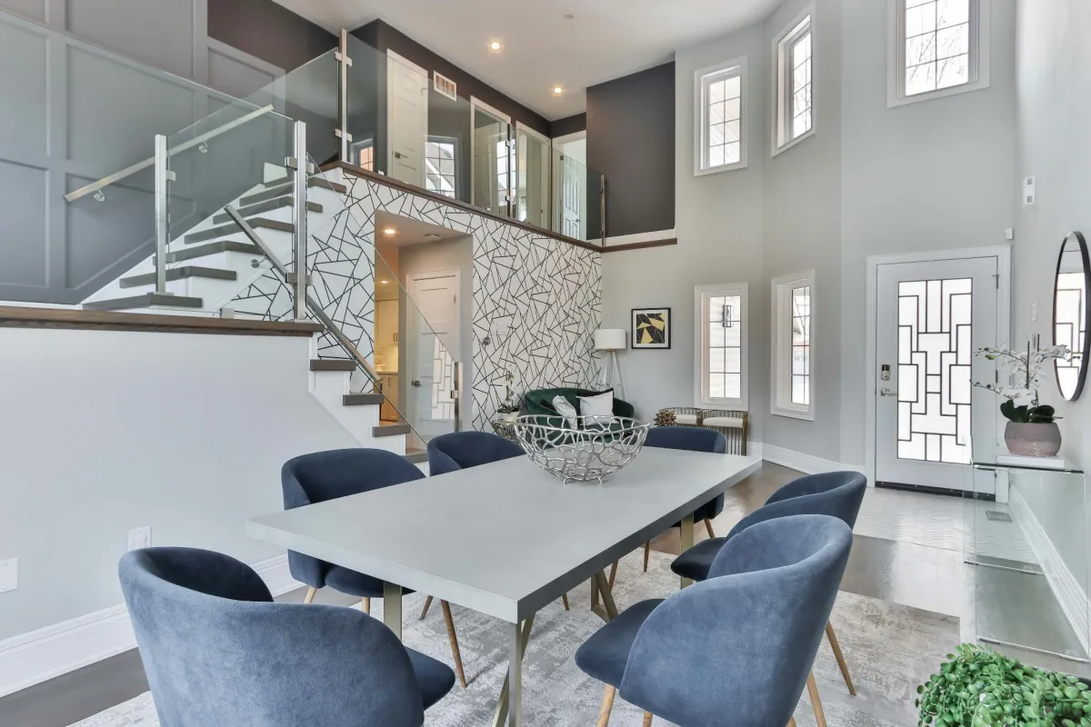 A modern dining room with a sleek table, blue velvet chairs, and a geometric accent wall near a glass staircase.