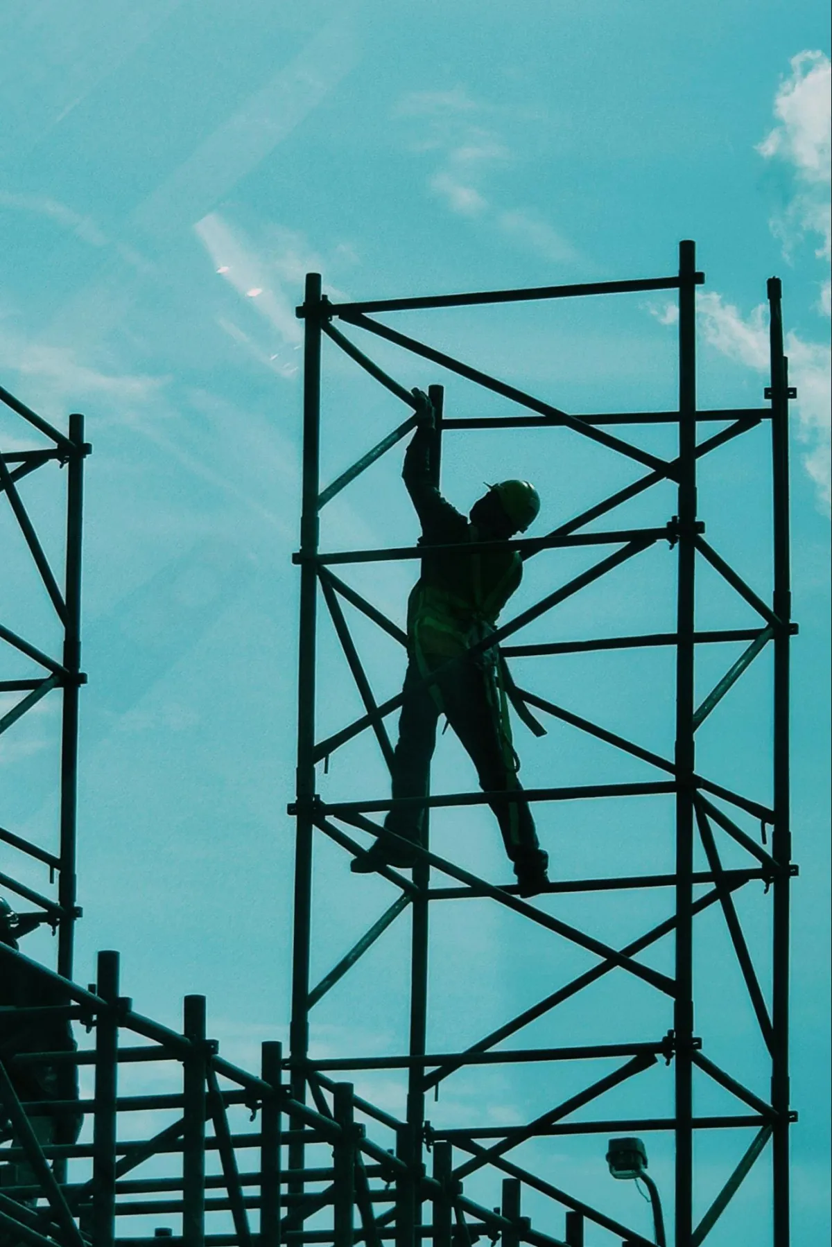 A construction worker wearing a safety harness and helmet climbs a metal scaffolding structure against a bright blue sky