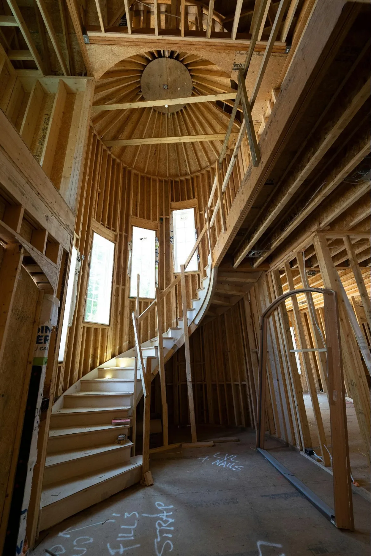 A partially constructed interior of a house featuring a curved wooden staircase, exposed framing, and a circular ceiling structure with large windows