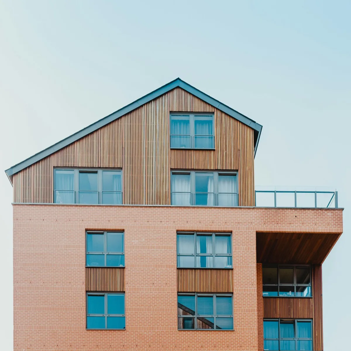 Close-up of a modern custom home with a combination of brick and wooden siding, featuring large windows and a rooftop terrace, under a clear blue sky.