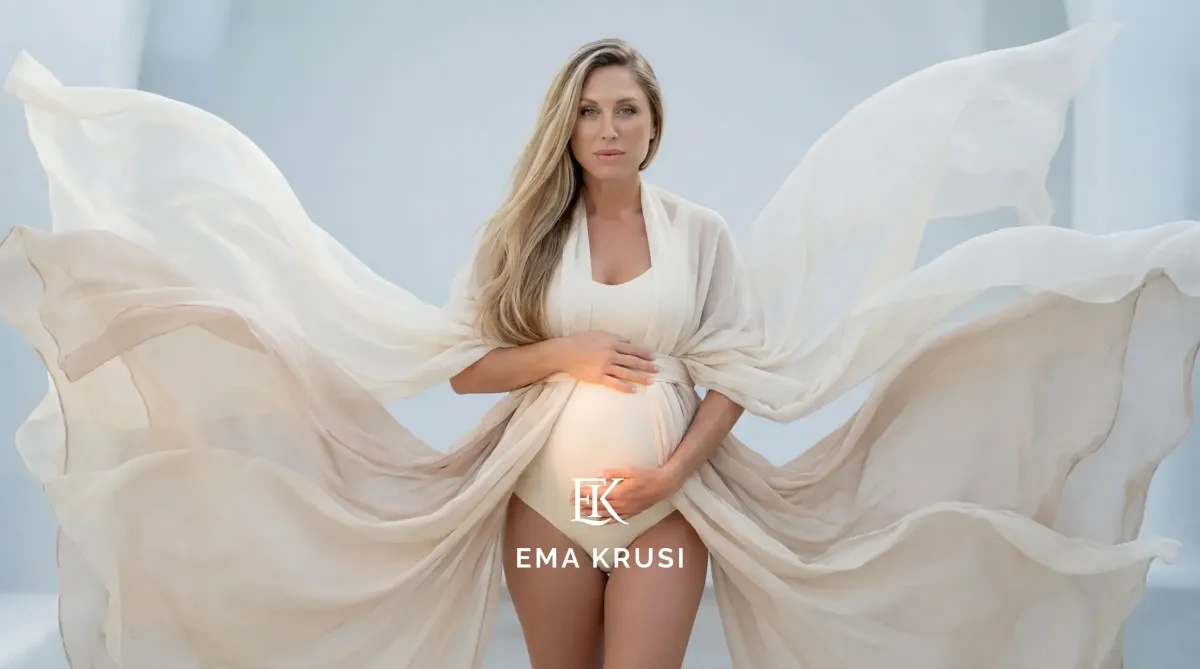 Portrait de femme enceinte en body clair, enveloppée d’un voile beige flottant, sur fond bleu doux.