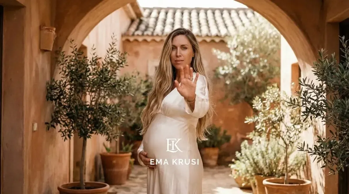 Femme enceinte en robe blanche levant la main pour dire stop, dans une cour aux murs terracotta et plantes en pots.