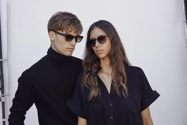 Fashionable couple wearing sunglasses, posing together with neutral background, one man in a black turtleneck and one woman in a black shirt.