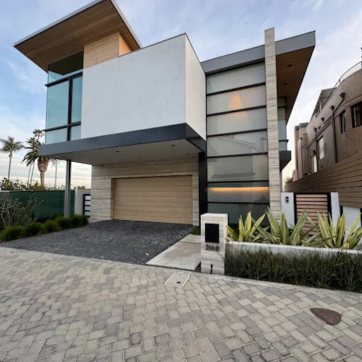 Modern two-story home featuring white stucco, extensive black-framed glass windows, a light wood garage door, and paving stone driveway.