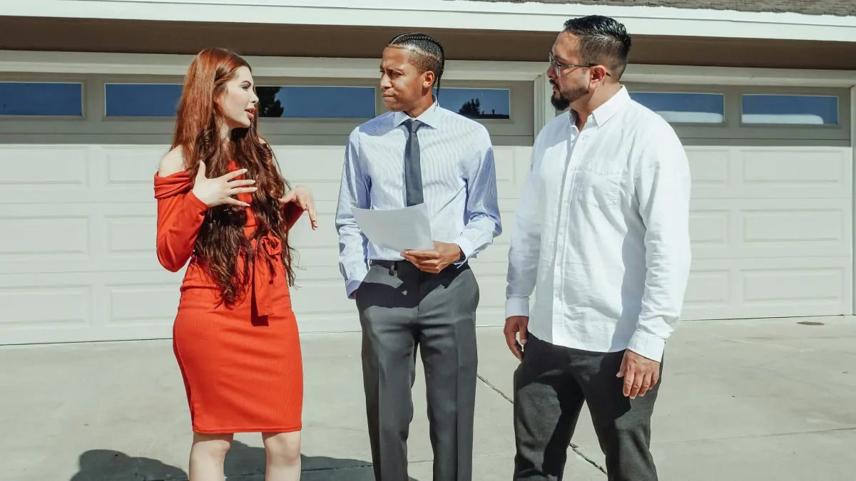 Outdoor meeting between three adults (two male, one female) standing on a driveway in front of a modern suburban home with a closed white garage door.
