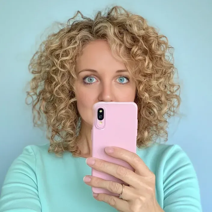 woman with curly hair, holding a pink smart phone upto her face, pale blule background and pale mint greenm top