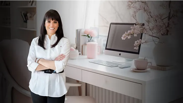 Femme souriante en chemise blanche, bras croisés, debout devant un bureau élégant et féminin avec un ordinateur de bureau, des fleurs et des accessoires roses, dans un espace de travail lumineux et inspirant.