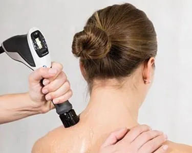 A therapist applying shockwave therapy to a woman's upper back using a handheld device. The woman has her hair in a bun and is receiving treatment for pain relief or muscle recovery