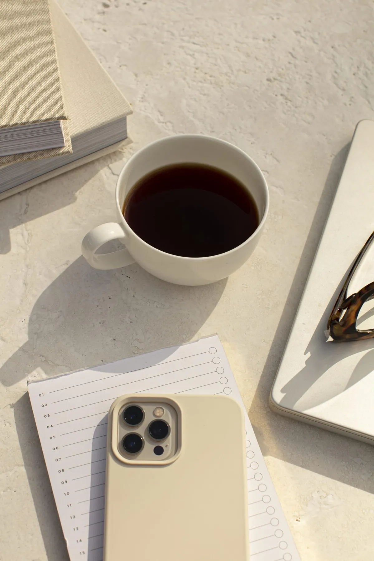 A desk scape in neutral cream colors featuring a cup of coffee, a notebook, a pair of glasses, and a cell phone.