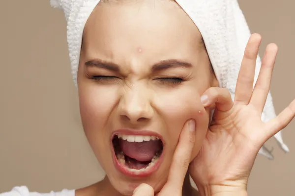 Person with a towel on their head, squeezing a pimple on their cheek, eyes closed, and mouth open.