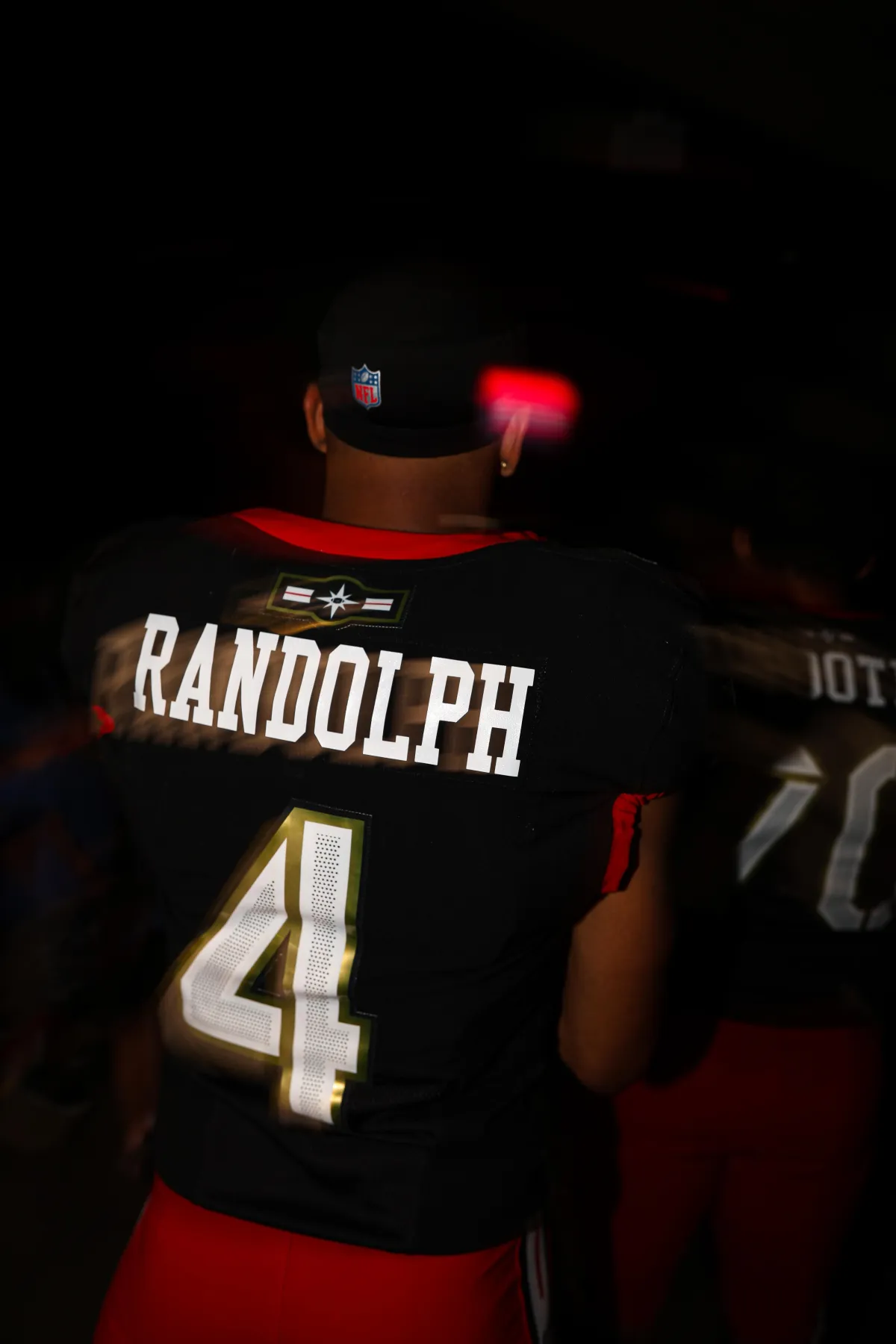 Football player wearing a black jersey with the name Randolph and number 4 in gold and white, standing in a dark tunnel with a red glow behind him.