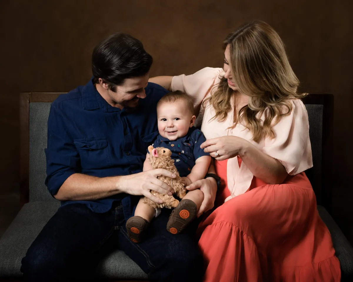 A smiling baby sits between two parents on a dark bench, holding a small stuffed animal. The father, wearing a navy shirt, and the mother, wearing a blush and coral dress, look lovingly at their child, creating a warm and joyful family portrait against a dark brown background.