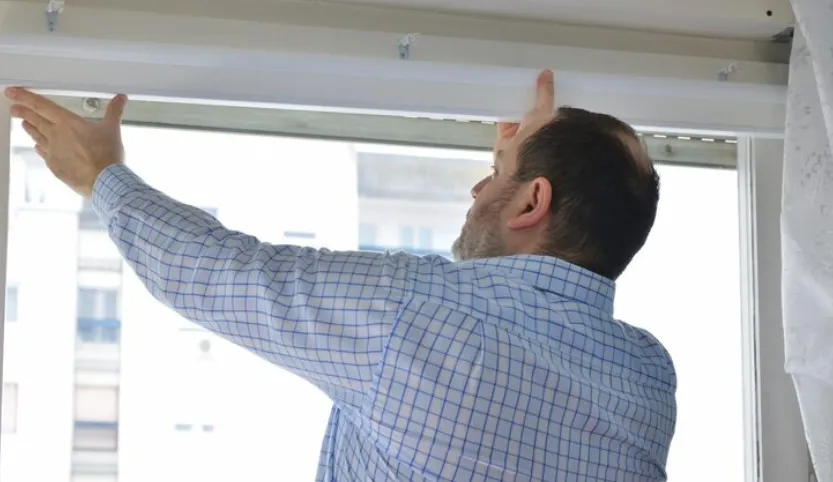 Person in a blue checkered shirt installing a window blind or curtain rod, reaching up to secure it above a window with a city view and white curtain.