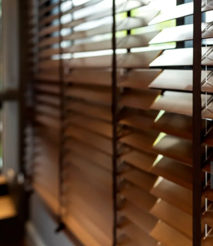 Close-up of horizontal wooden window shutters with evenly spaced slats, partially open to let in filtered light.
