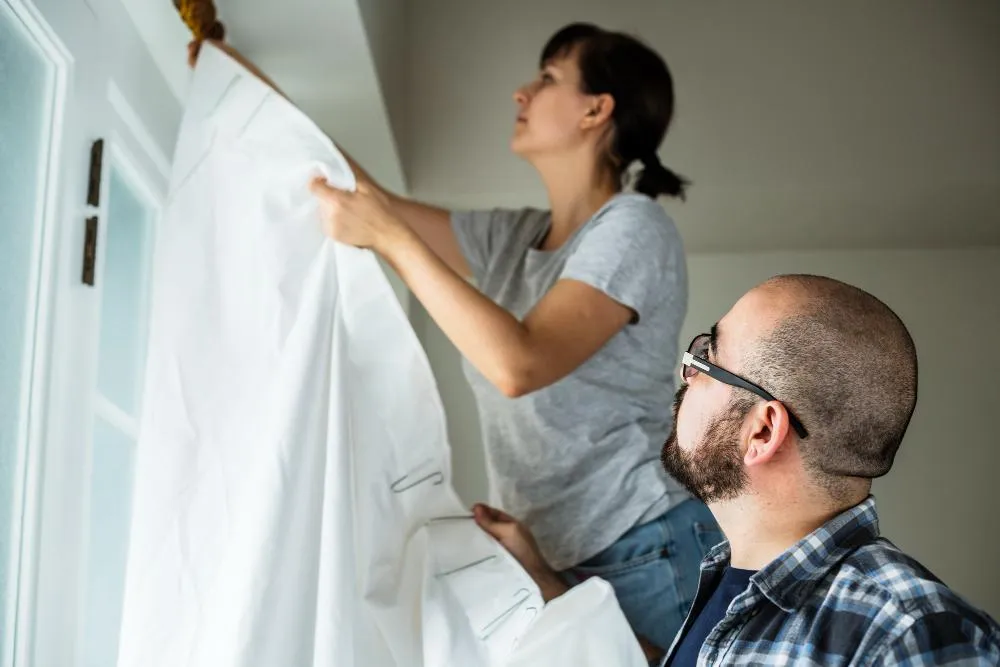 Woman and man working together to remove curtains from a window, preparing the space for new window treatments.