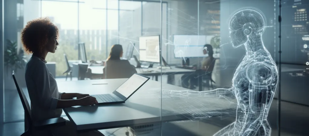 A businesswoman works on a laptop in a modern glass-walled office while a transparent holographic humanoid AI figure sits across from her, symbolizing collaboration between humans and artificial intelligence. Other employees are visible working at desks in the background.