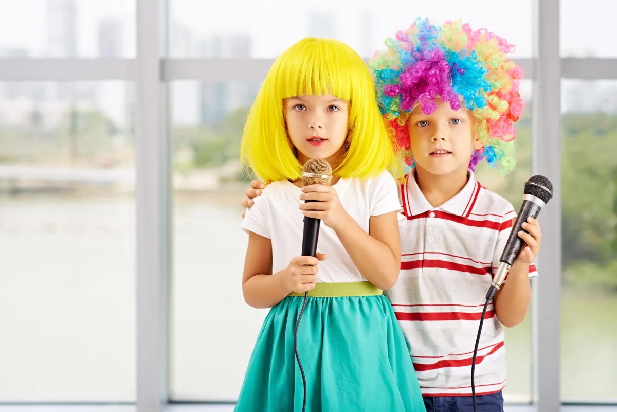 The image shows two young children dressed up in colorful wigs, each holding a microphone as if they are about to perform or sing. The child on the left is wearing a bright yellow bob-style wig, a white shirt, and a turquoise skirt. The child on the right is wearing a rainbow-colored curly wig and a striped polo shirt. They are standing close together with their arms around each other, and they appear to be having fun, possibly engaged in a playful performance or karaoke. The background suggests they are indoors, with large windows allowing natural light in. The overall mood is lighthearted and playful.