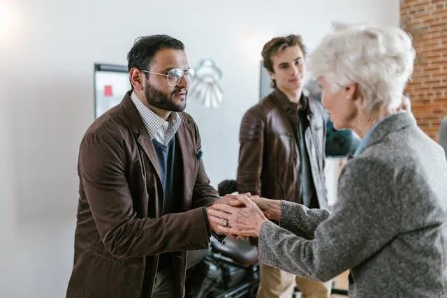 The photo depicts a caregiver and a support worker engaging with an elderly woman. The caregiver, a middle-aged man wearing glasses and a brown jacket, is shaking hands with the elderly woman, showing a gesture of support and assurance. In the background, a younger man observes the interaction, indicating a team or family setting. This image effectively illustrates a welcoming and professional introduction, suitable for a blog about onboarding support workers in a caregiving environment.