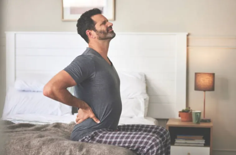 A mature man feeling back pain at home while sitting on his bed after waking up from being asleep.