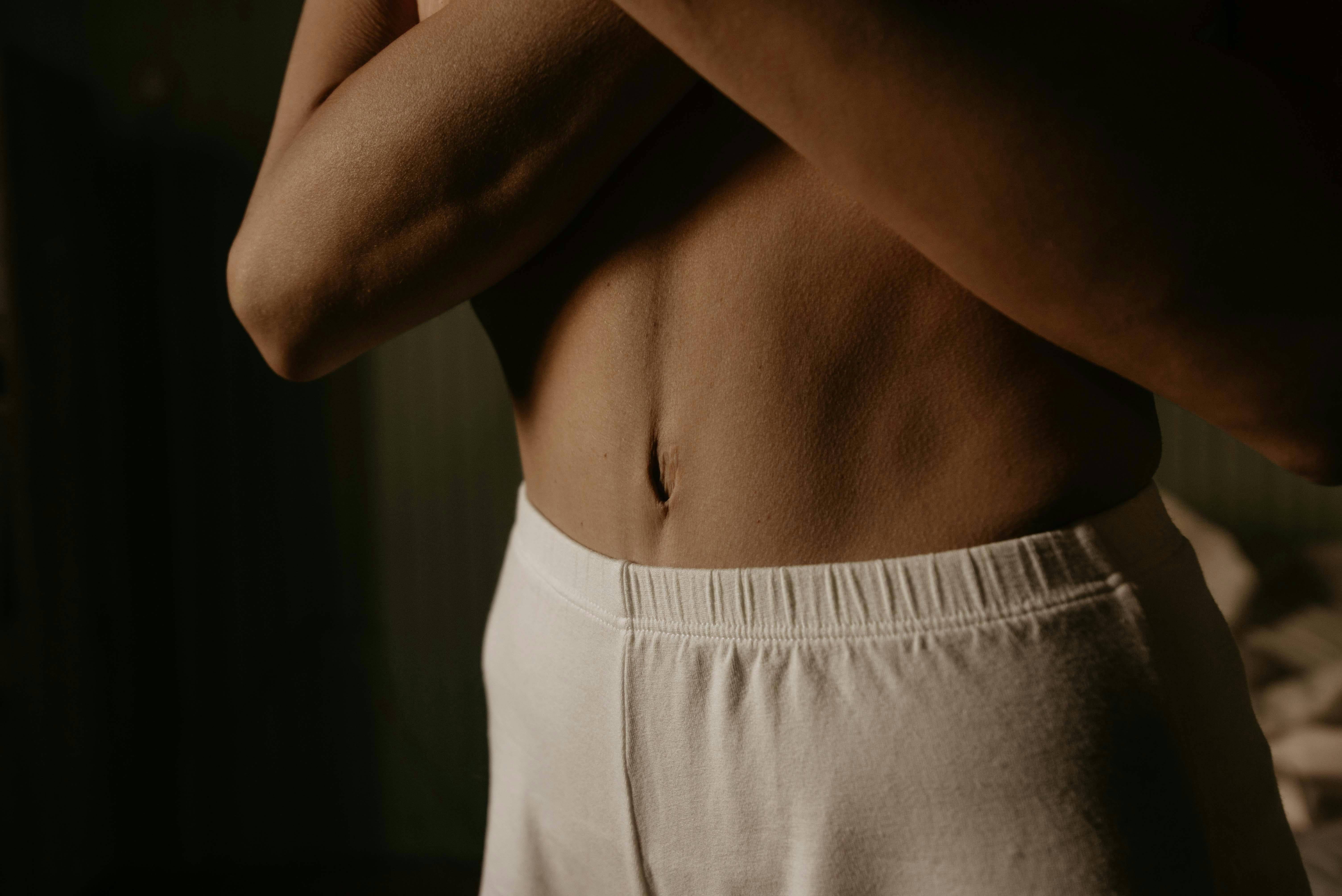 Close-up of a fit adult male torso with defined abs, wearing light-colored shorts in a soft, moody setting, representing men’s health, confidence, and physical well-being.
