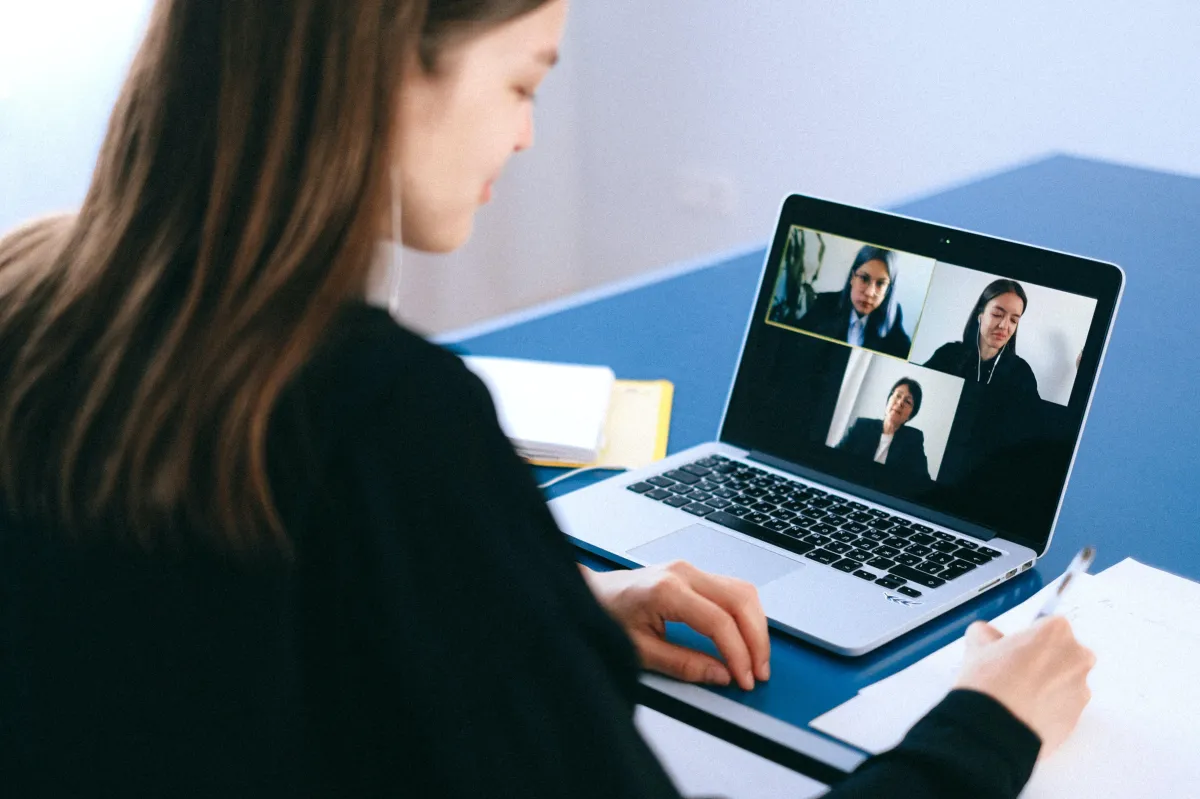 Woman taking notes in a notebook while talking to 3 other people in a video conference.