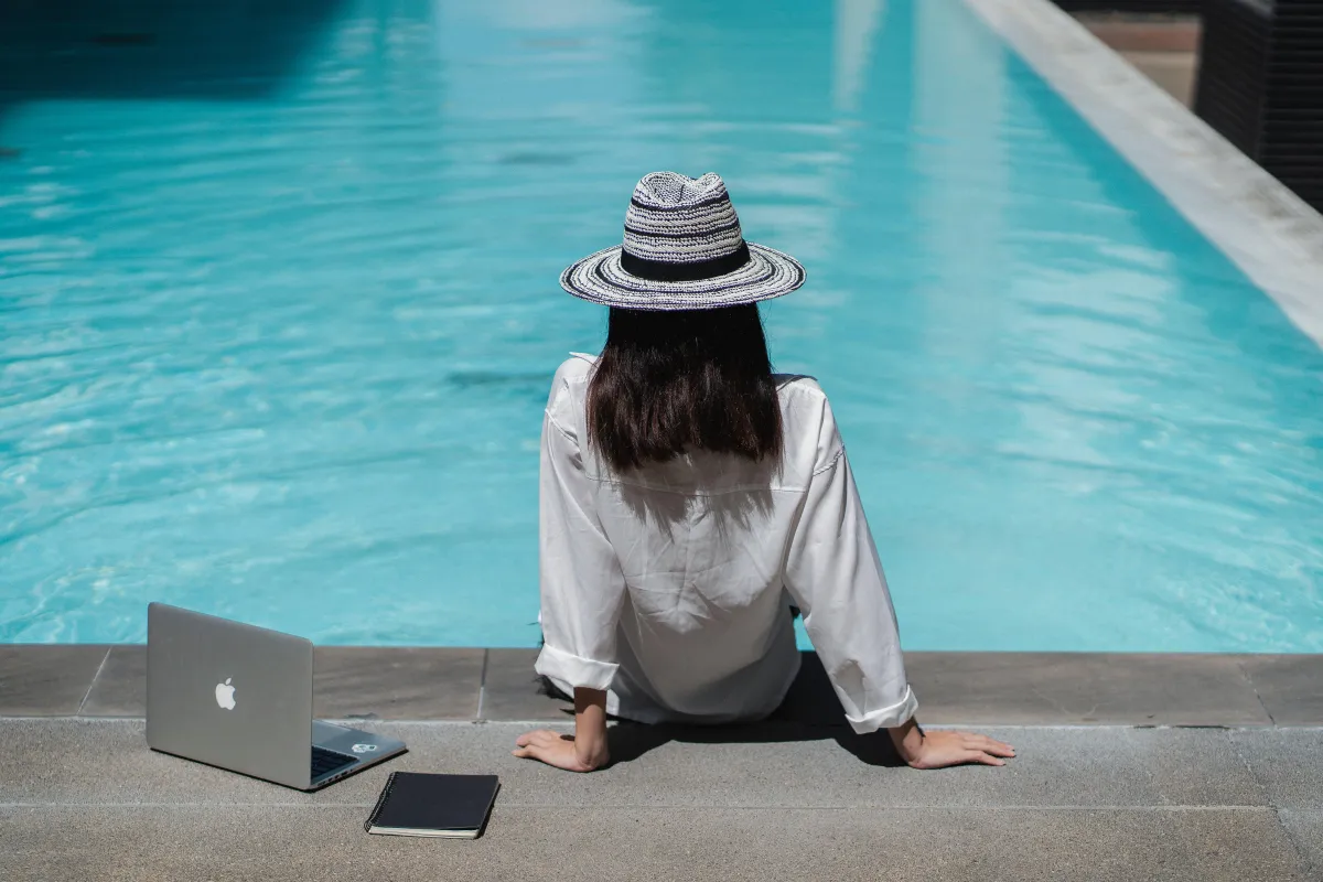 Person sitting at the edge of a pool with legs in the water while their work laptop is open next to them