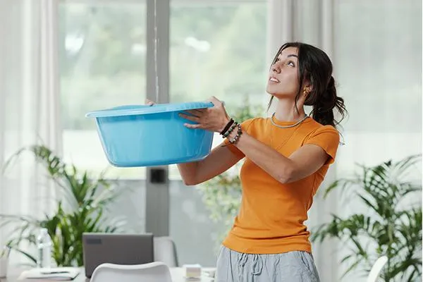 Concerned woman holding a blue bucket indoors, looking up at the ceiling, indicating a roof leak issue in a bright living space with plants.