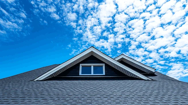 A modern house with a dark gray asphalt shingle roof and white-trimmed gables against a bright blue sky with scattered white clouds.