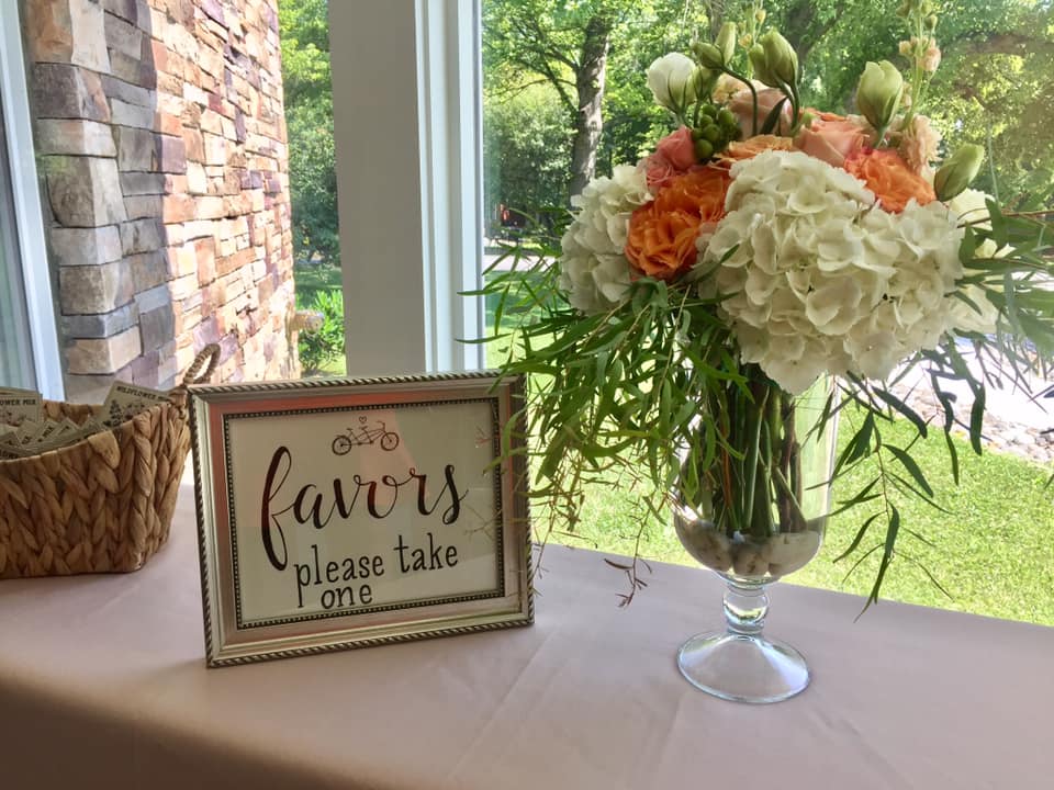 Favors table floral arrangement in a tall vase with white hydrangea and orange Free Spirit roses.
