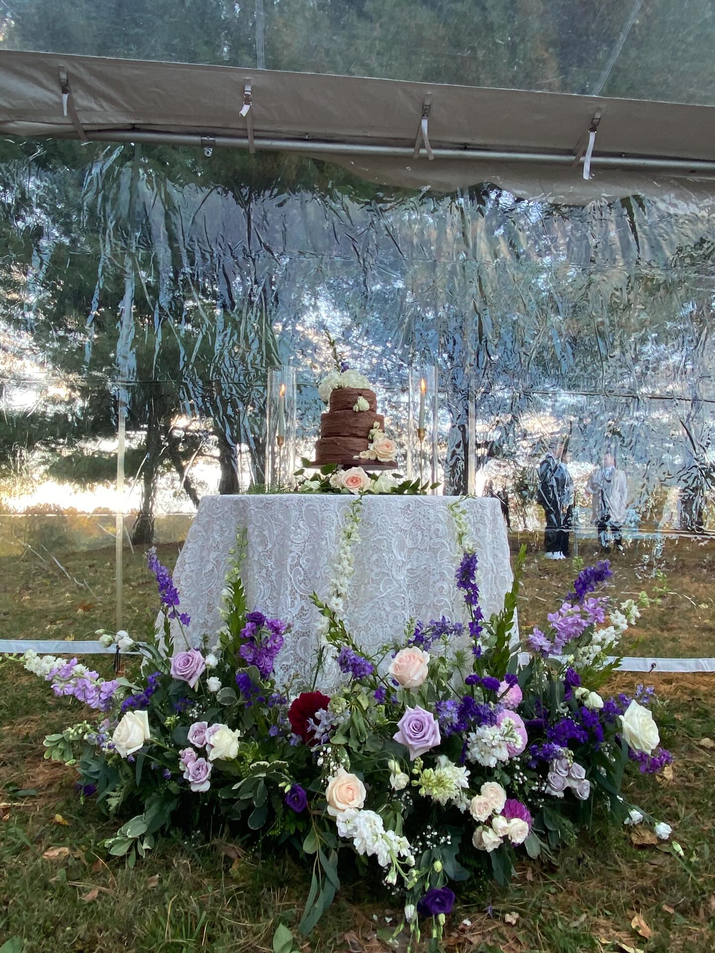Seasonal wedding floral installation surrounding a cake table with roses, delphinium, and greenery under a clear tent