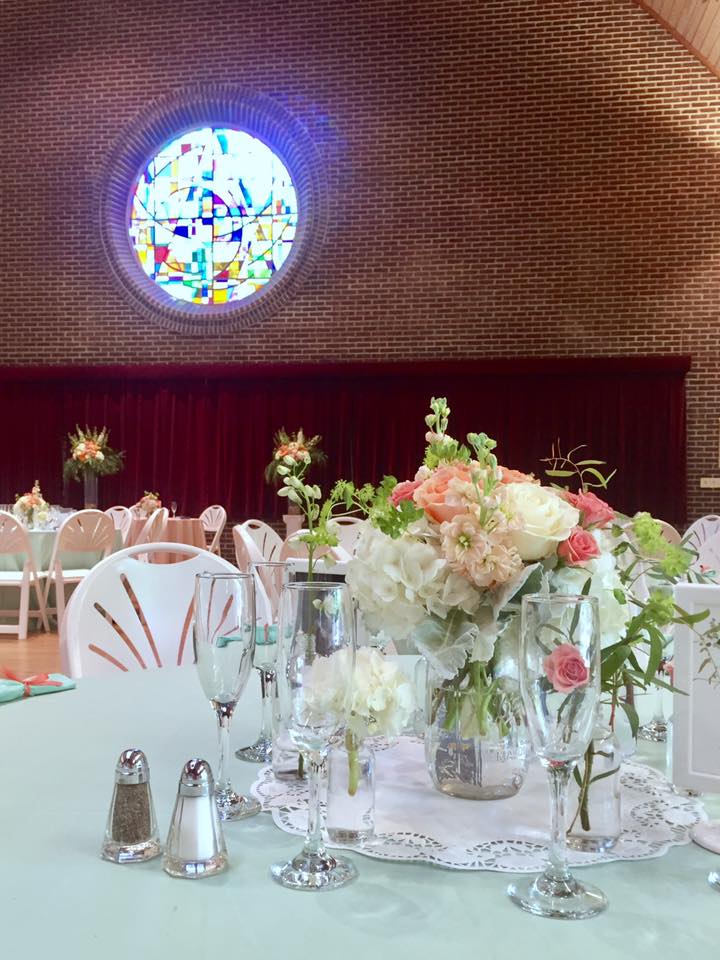 Wedding reception room with round tables set with mint green linens and floral centerpieces in creamy white, orange, peach, and pink.