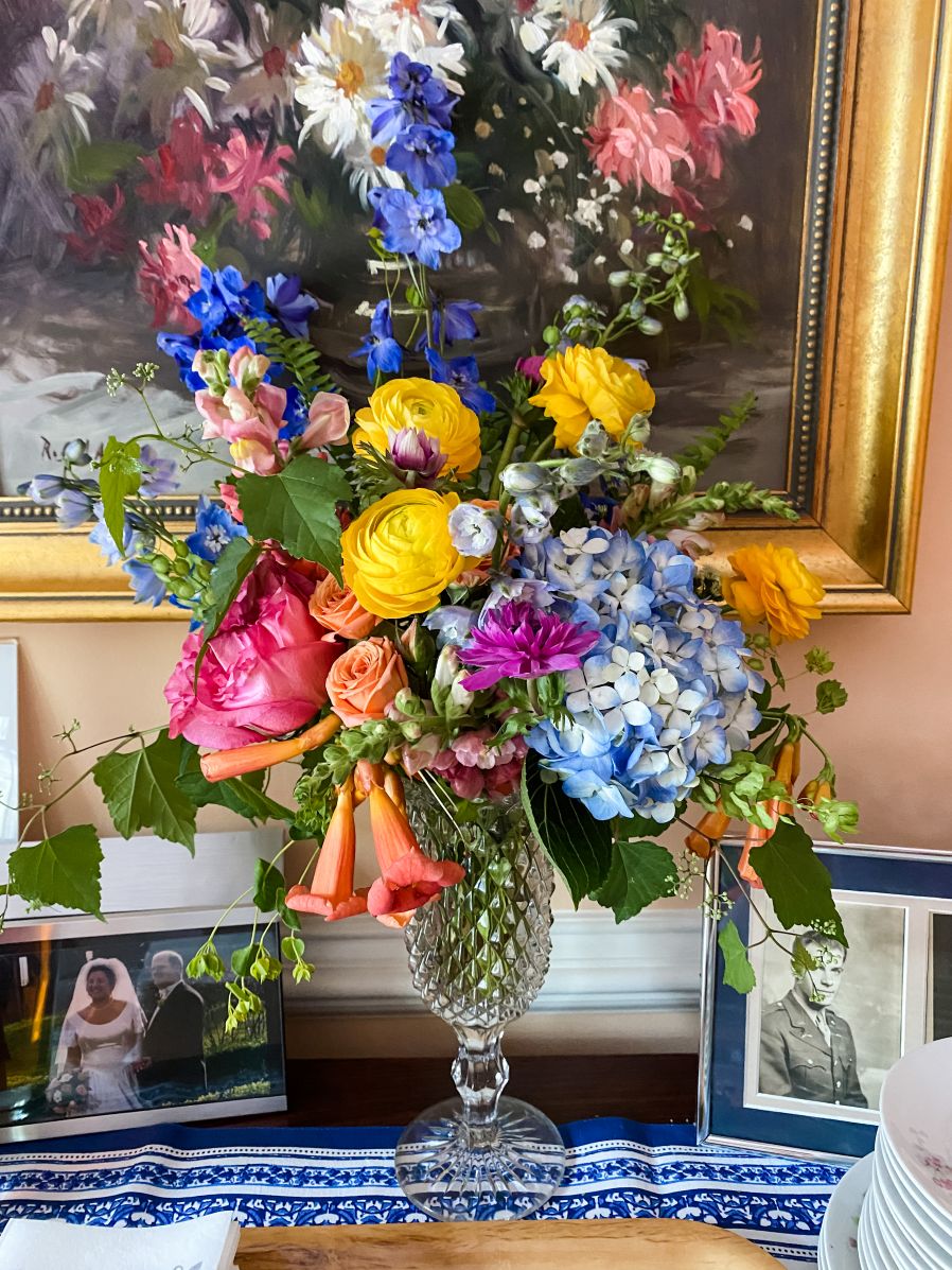 Buffet Centerpiece in vintage glass footed vase with hot pink peony, blue hydrangea, yellow ranunculus, cobalt blue delhinium, orange spray roses, orange snapdragons, and orange trumped flower.