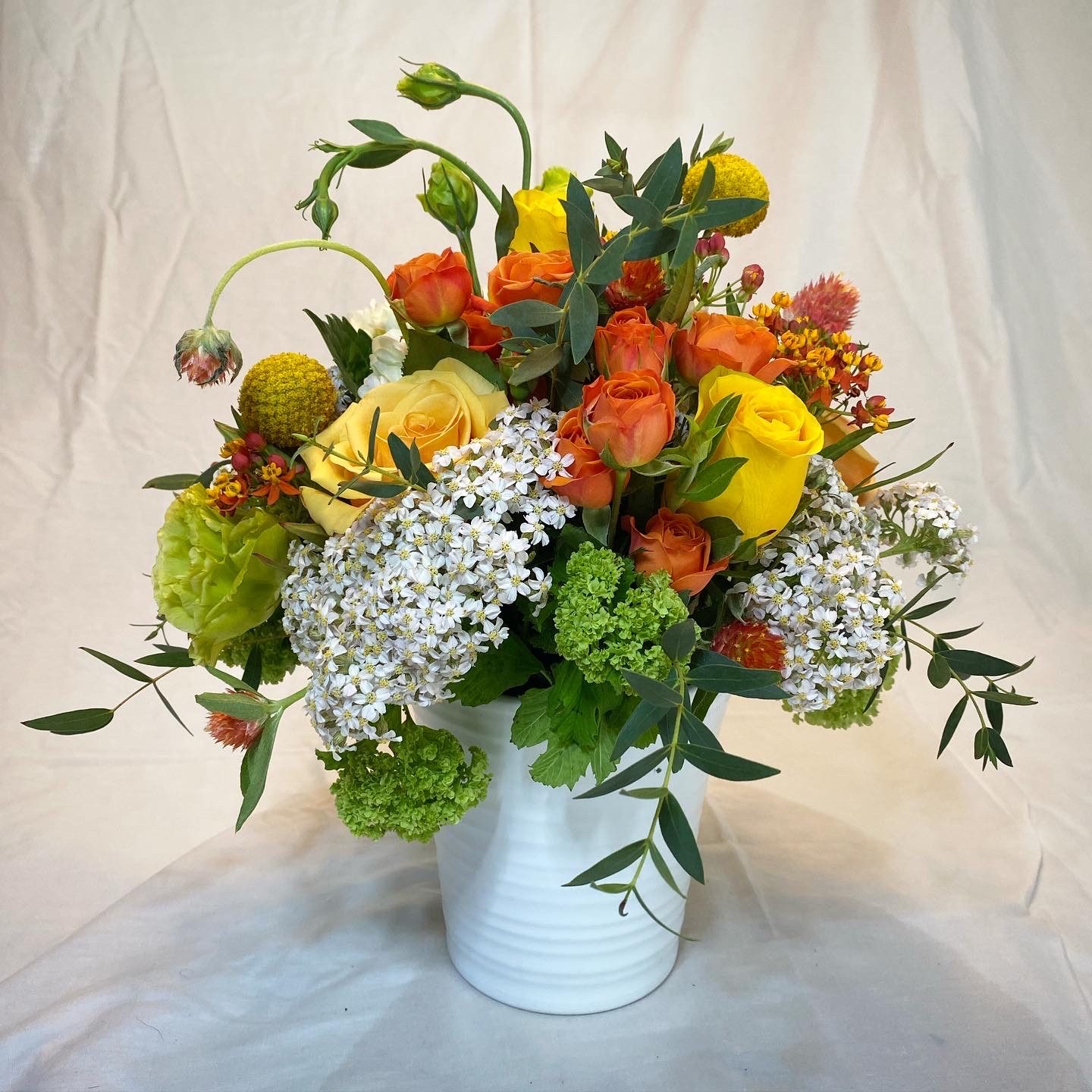 Centerpiece with yellow roses, orange spray roses, gomphrena, craspedia, lisiantus, viburnum, yarrow, and eucalyptus