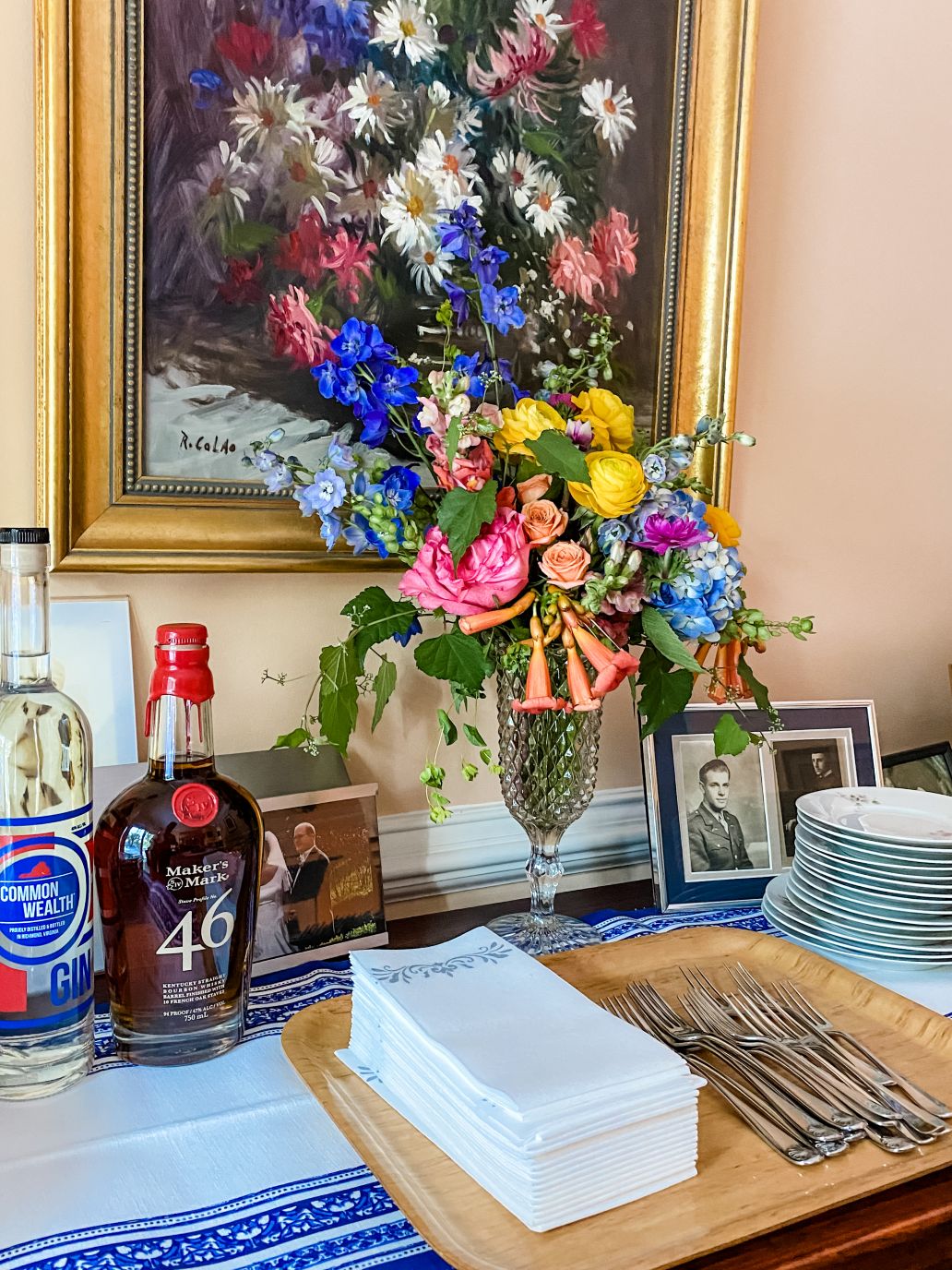 Tall, colorful buffet centerpiece with dark blue delphinium, light blue hydrangea, hot pink peonies, orange spray roses, yellow ranunculus, and orange trumpet flower vine