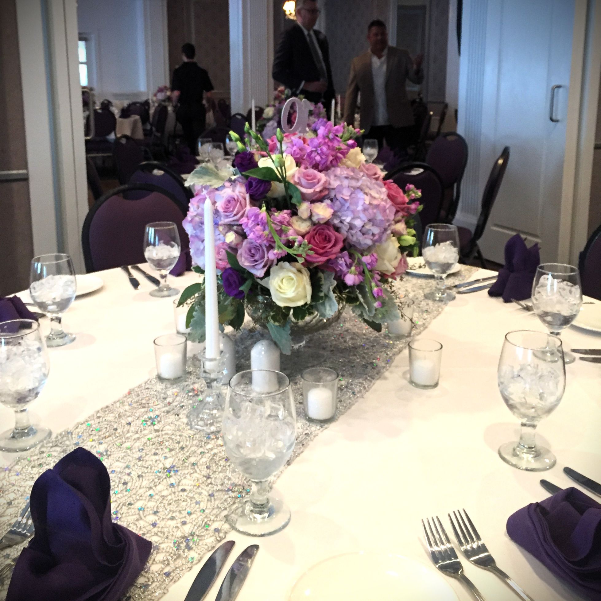 Dining table centerpiece in shades of purple, cream, and lavender with roses, spray roses, hydrangea, lisianthus, dusty miller, and variegated pittosporum