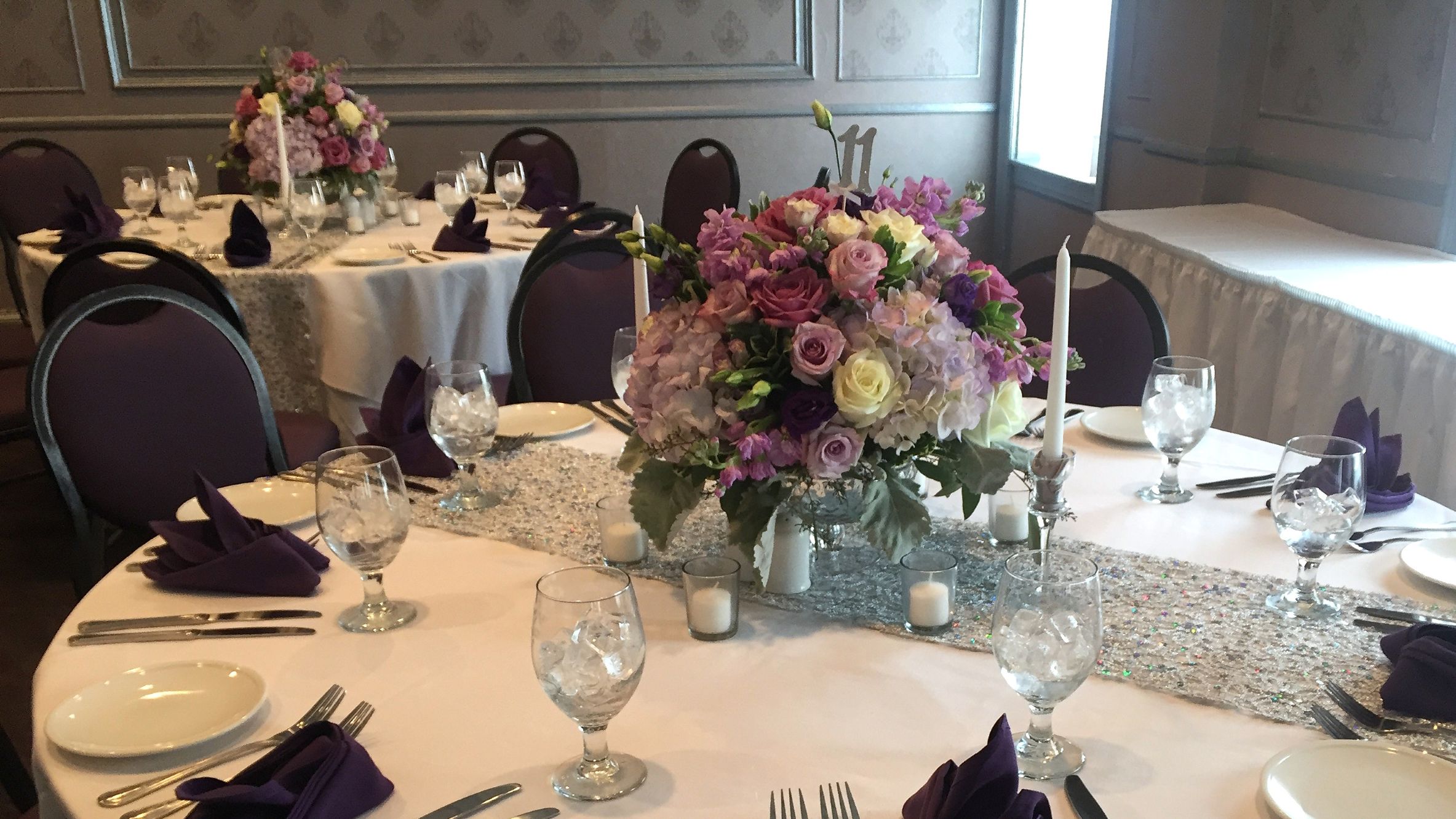 Reception dining tables with large floral centerpieces in shades of purple, lavender, and white.