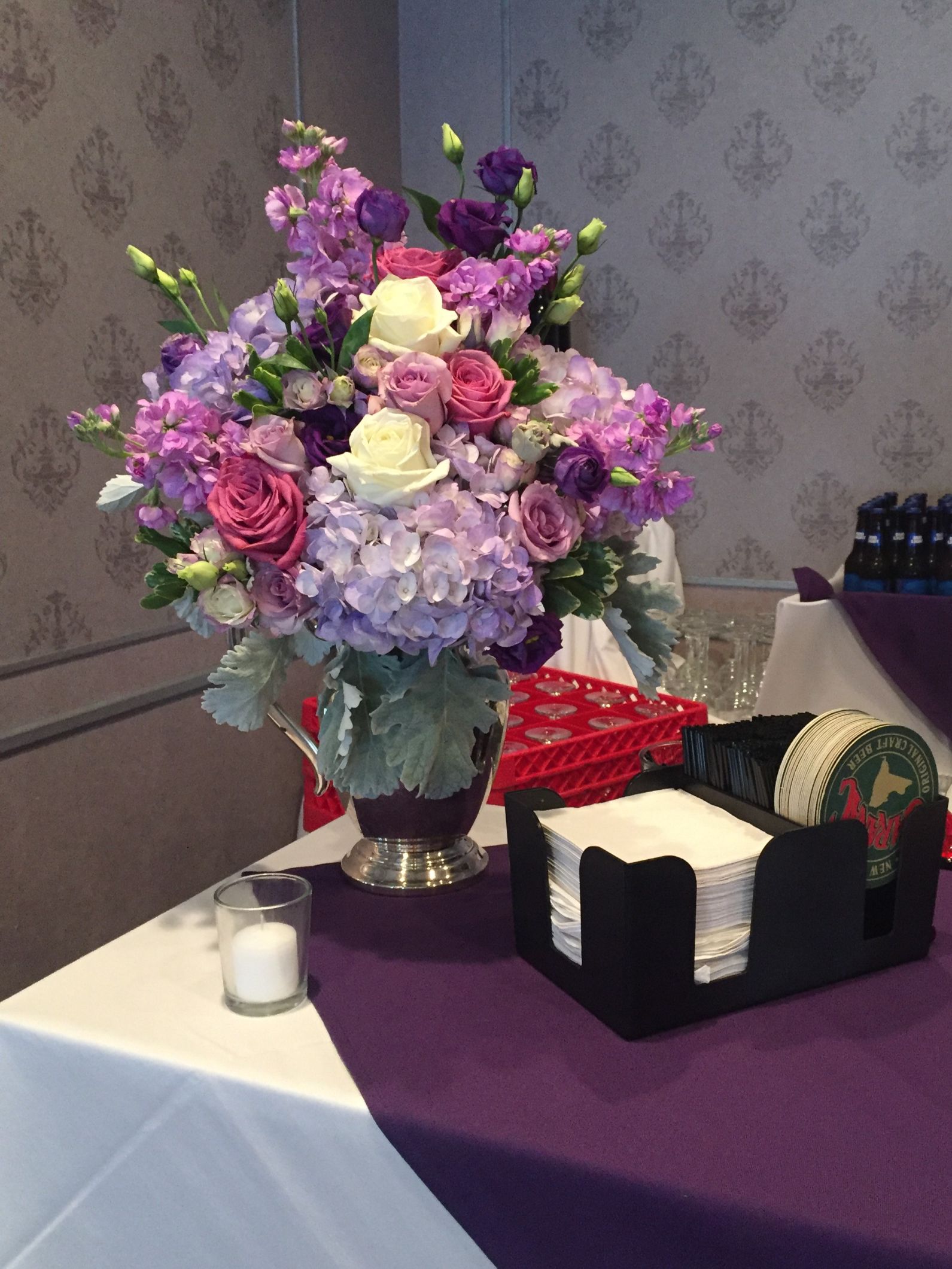 Bar floral in shades of purple with hydrangea, roses, stock and lisianthus in a vintage silver pitcher 