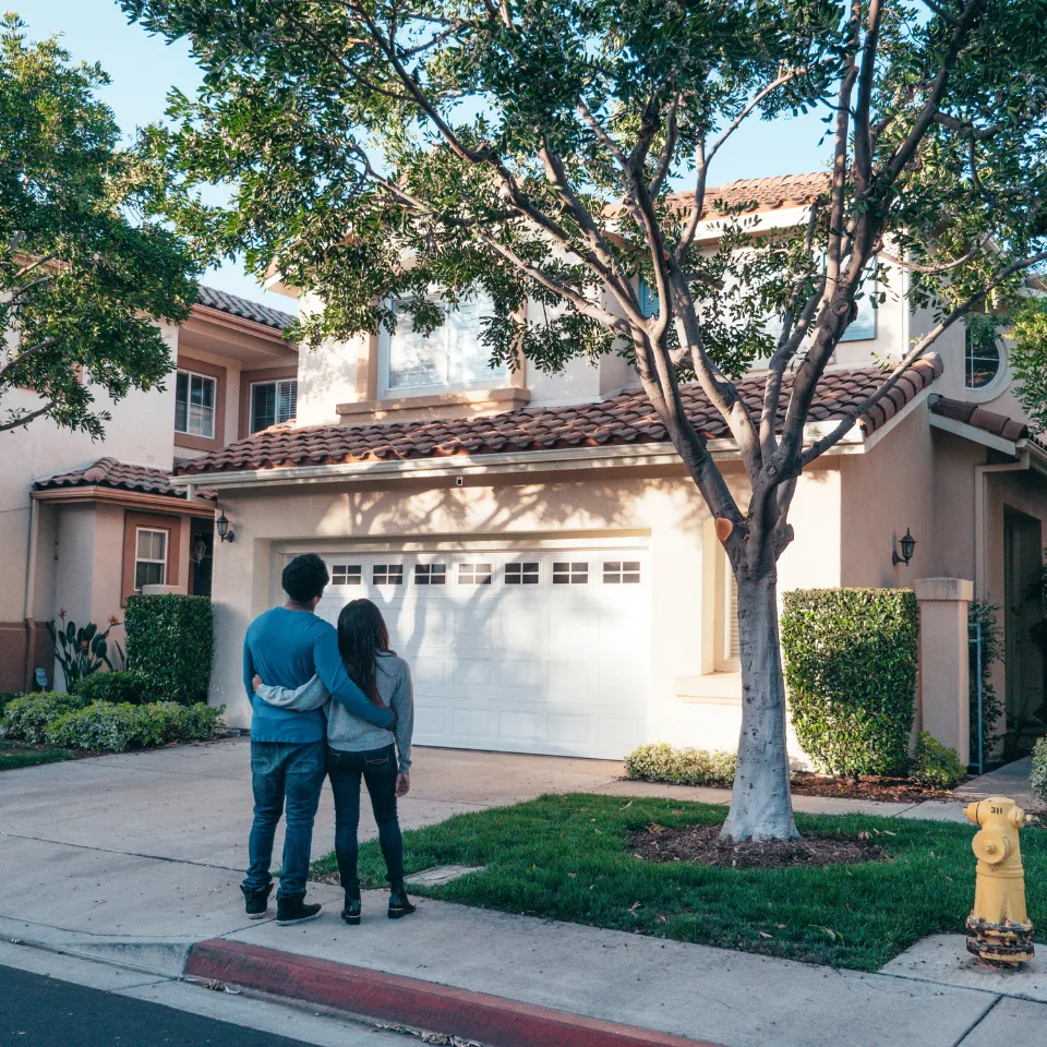 Happy first-time homebuyers standing in front of a new house holding keys and paperwork, with a real estate agent nearby.