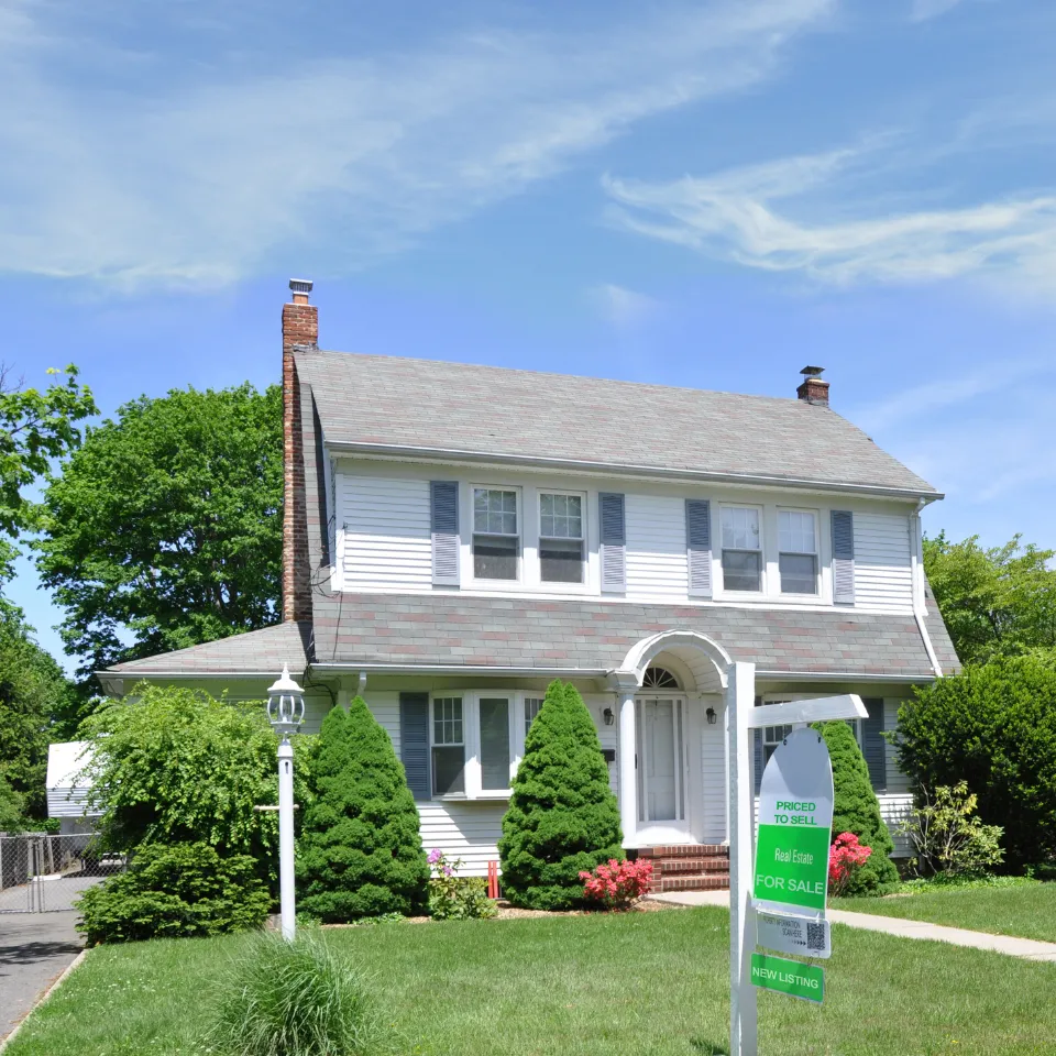 Modern home with blooming spring flowers, clear skies, and a “For Sale” sign out front.