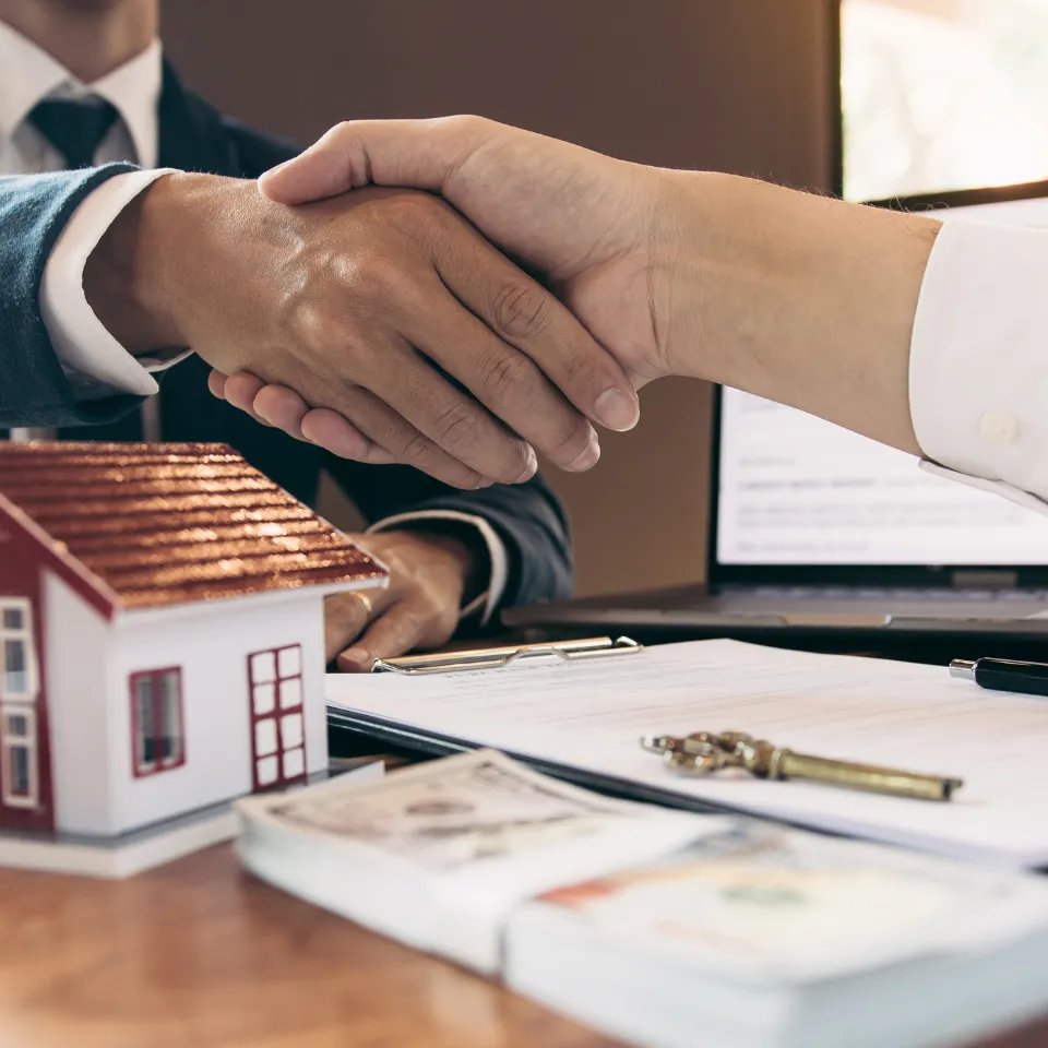Homebuyer and agent shaking hands across a table with a house model and documents in view.