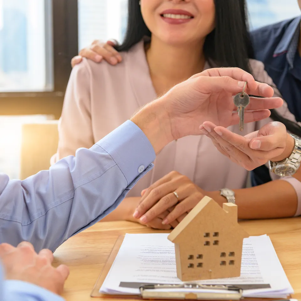 A smiling homeowner signing real estate documents with a "For Sale" sign in the background.
