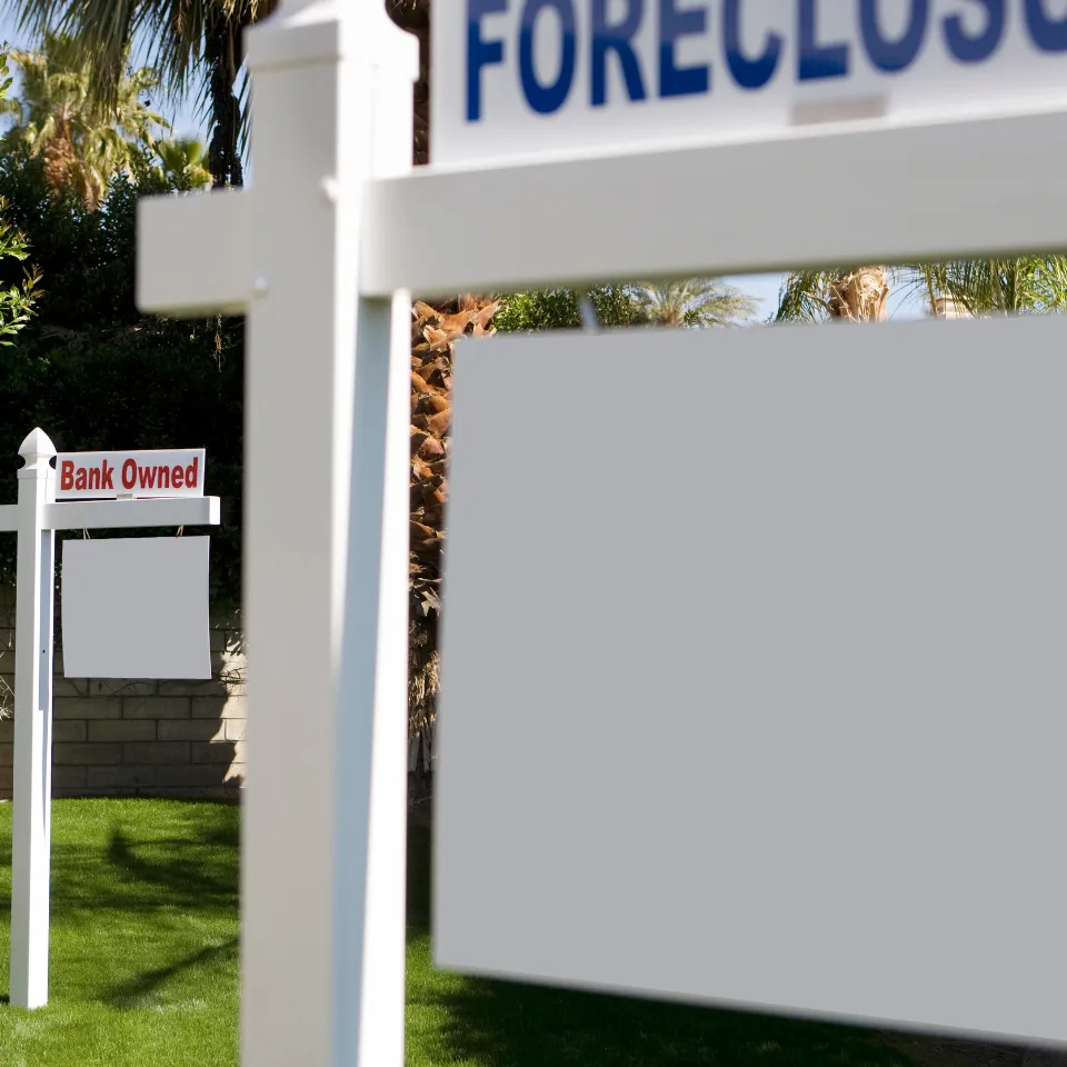 Exterior of a foreclosed home with overgrown lawn and "bank-owned" sign in front.