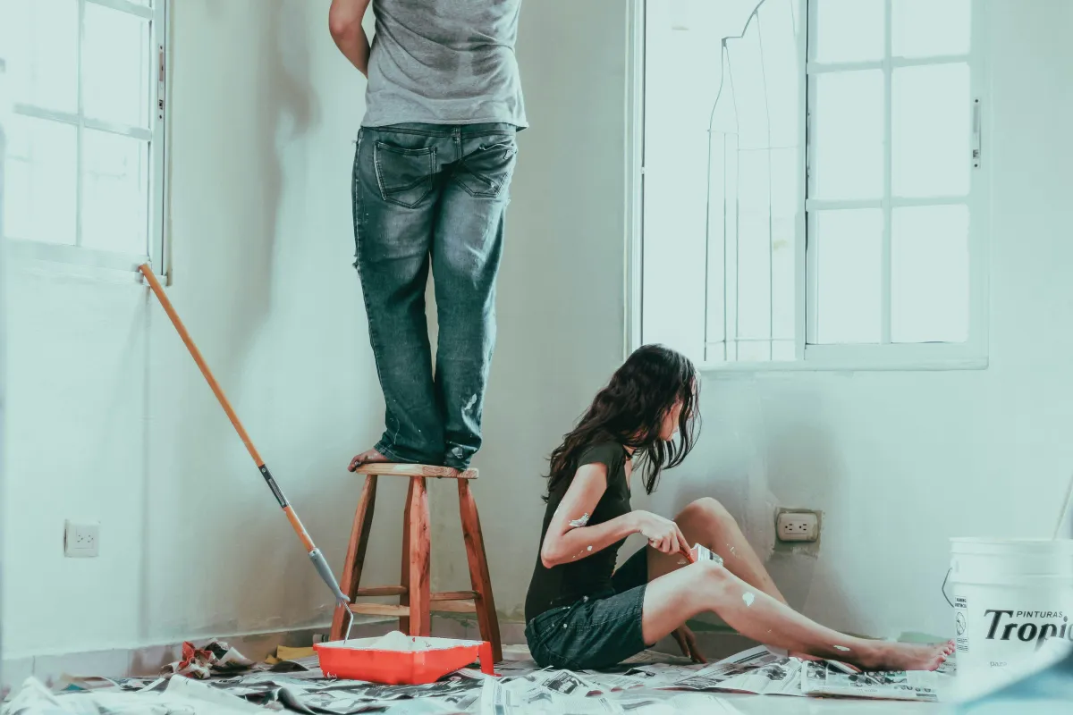 Couple painting a room: one stands on a stool, the other sits on newspaper-covered floor, with paint splatters and supplies.