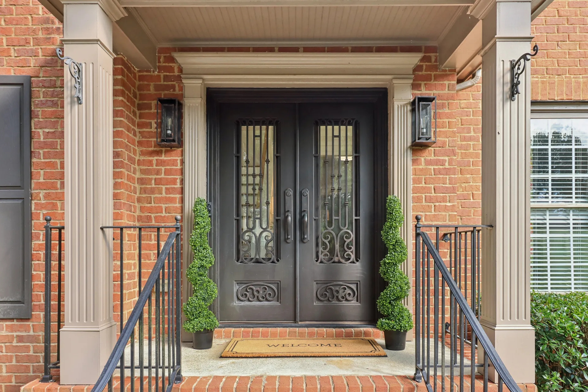 A high-resolution aerial photograph taken during golden hour, showcasing a vibrant suburban town center surrounded by lush green trees, upscale buildings, and a central fountain, with a serene lake and wooded residential neighborhoods extending into the background — representing the picturesque charm of North Atlanta suburbs like Johns Creek and Alpharetta.